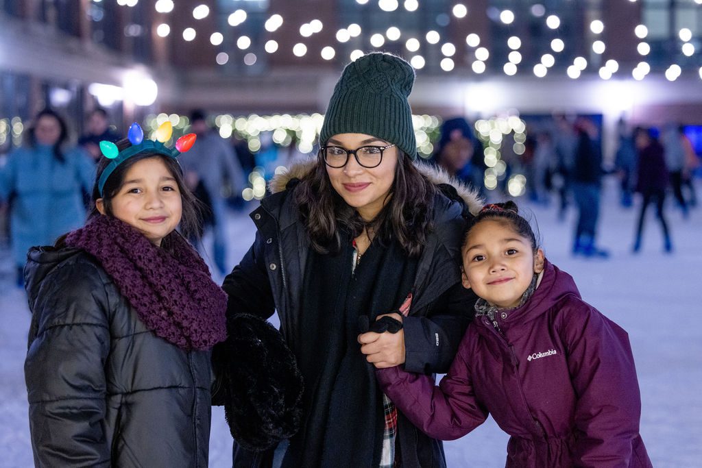 Mom in grey hat and glasses and two daughters in maroon on the ice skating smiling for photo
