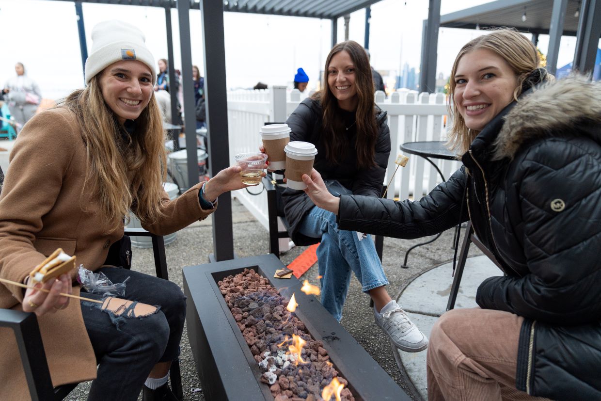 three women in winter coats toasting with drinks in hand sitting by the fire pit outside