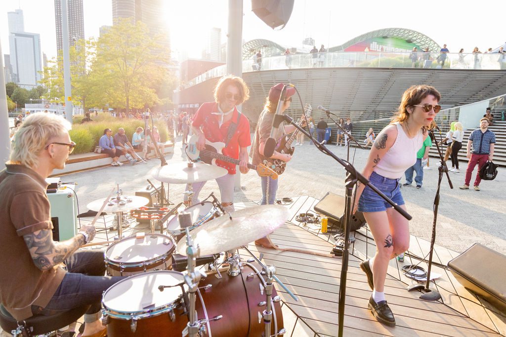 A group of musicians, including a singer, drummer, and guitarist, performing on an outdoor stage during summer.