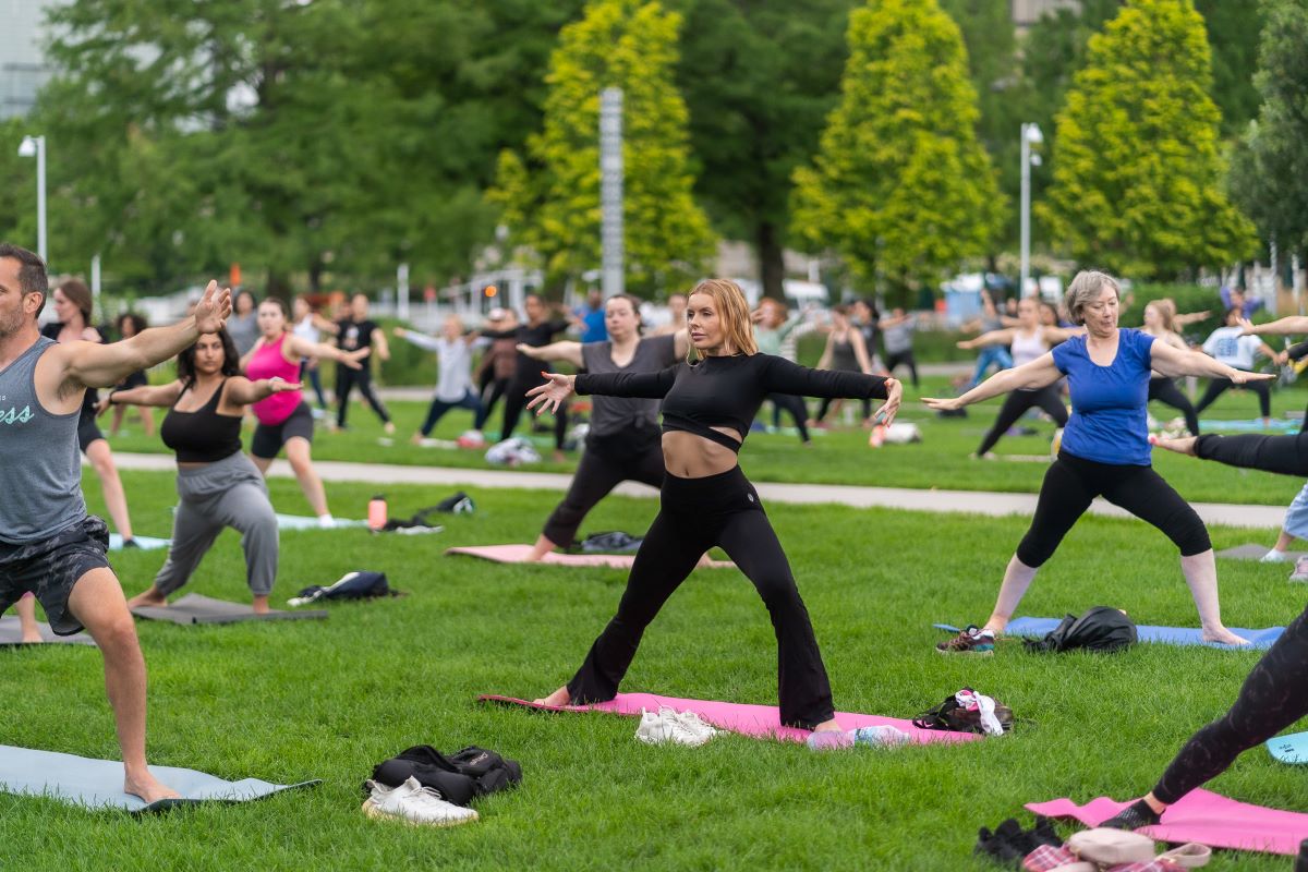 A group of people practicing yoga in a sunny park, surrounded by trees and grass, promoting wellness and relaxation