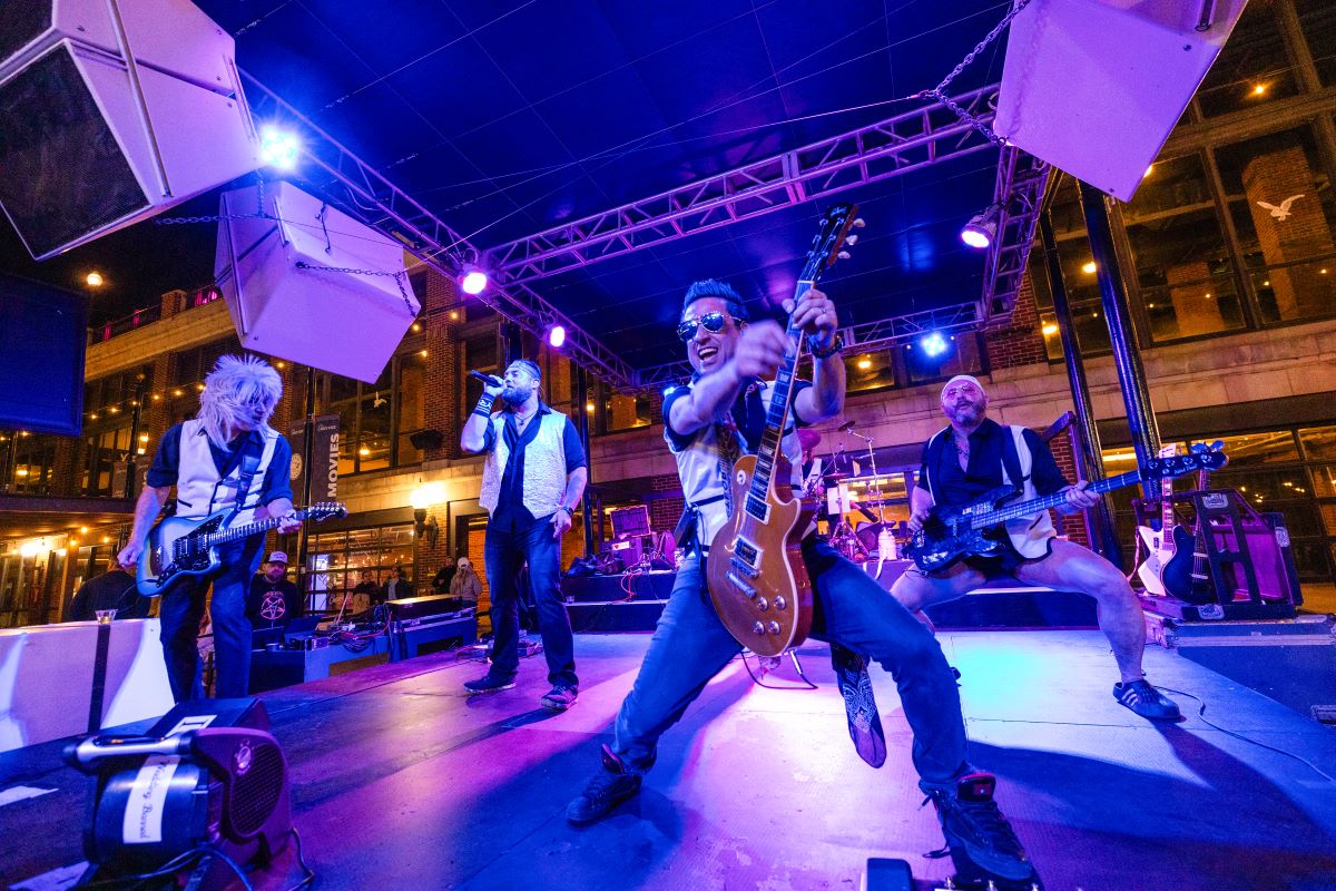 A band performs on an outdoor stage at night, illuminated by colorful lights during a summer concert.