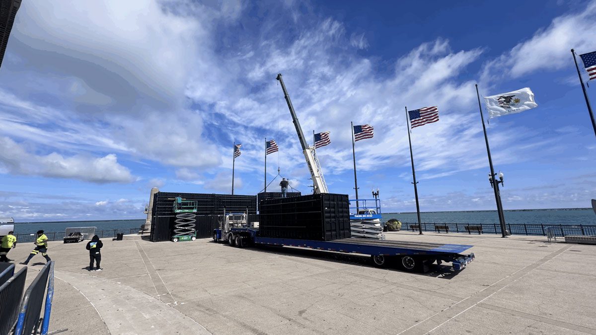 Workers preparing event staging and equipment at East End Plaza with American flags and lake backdrop