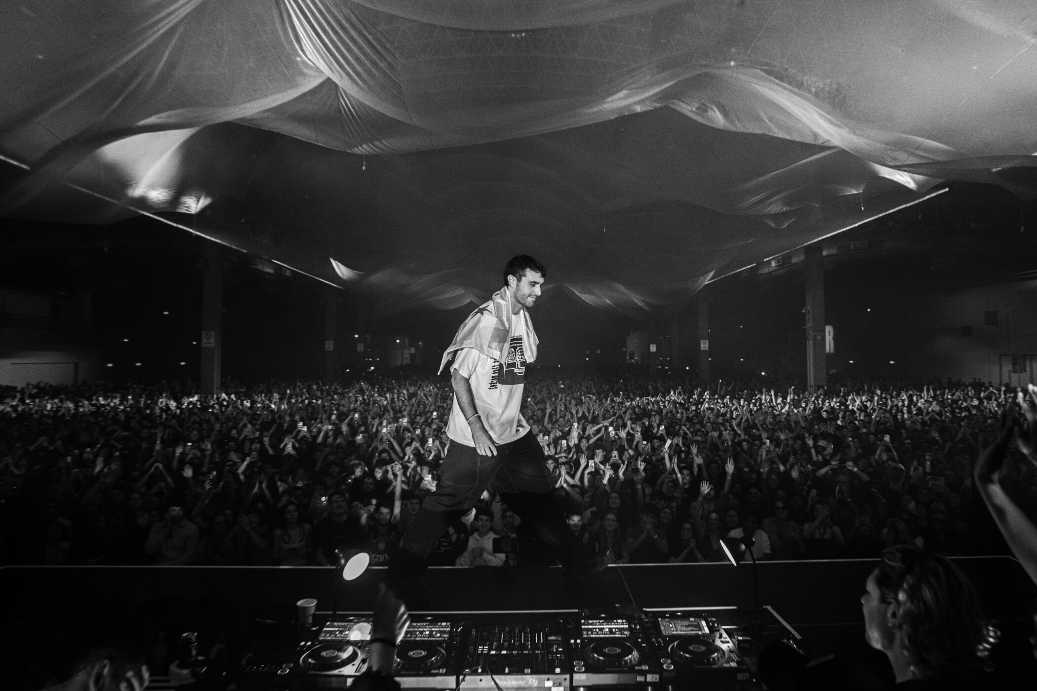 Black and white photo of fred again on stage facing a large indoor concert crowd